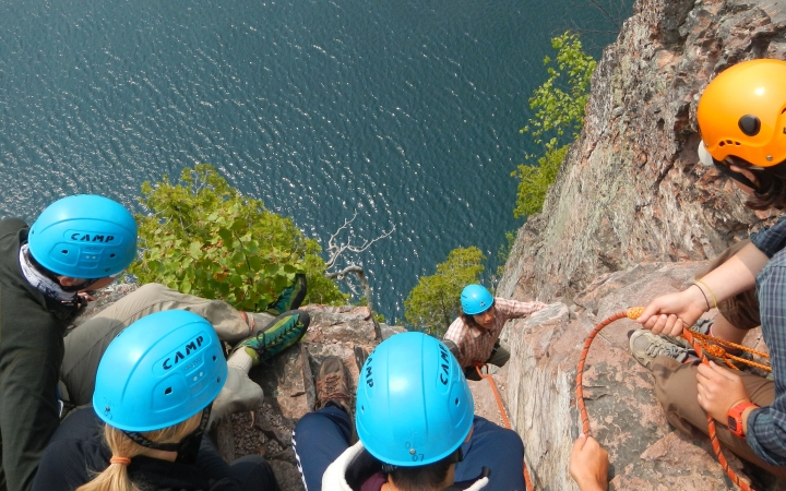 Four people wearing safety gear peer over the edge of a cliff, looking down at a rock climber making their way up. They are all above a blue body of water.